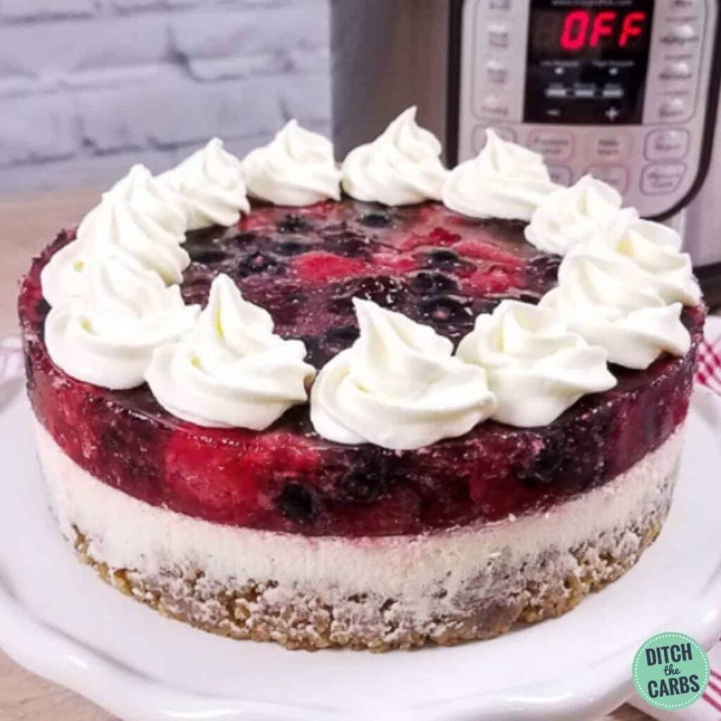 A close-up of a large slice of low-carb berry cheesecake on a white plate sitting in front of an Instant Pot and a read and white napkin