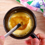 The back of a fork is pushing the lemon curd filling into the cake batter in a black mug.