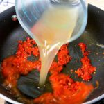 Pouring chicken broth out of a glass measuring cup into a black skillet that has diced red chili mixture in it.