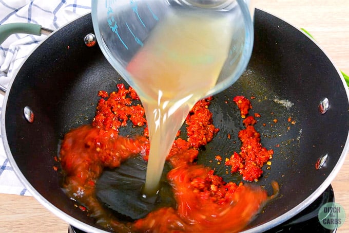 Pouring chicken broth out of a glass measuring cup into a black skillet that has diced red chili mixture in it.