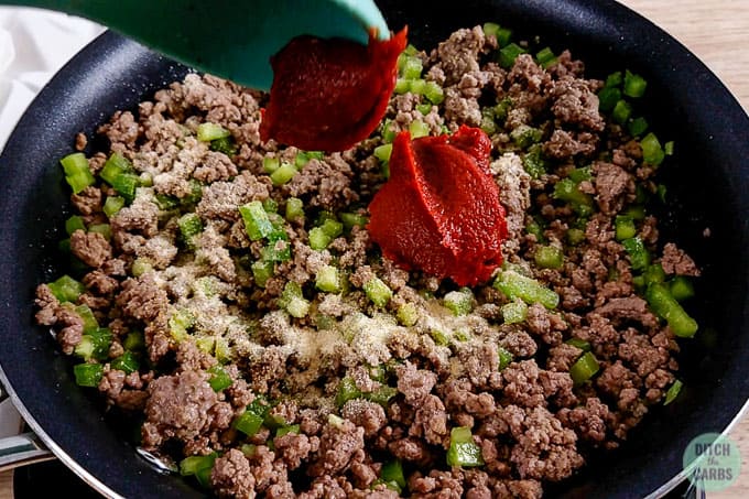 Adding seasoning and tomato paste to browned ground beef and chopped bell peppers in a skillet.