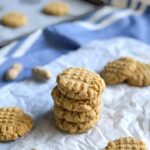 4 keto peanut butter cooked stacked on top of each other on a piece of white parchment paper. More cookies on laying off to the side and whole peanuts are scattered around the background.