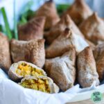 Keto samosas cooling in a dish lined with white parchment paper. One samosa has been cut in half to show the filling.