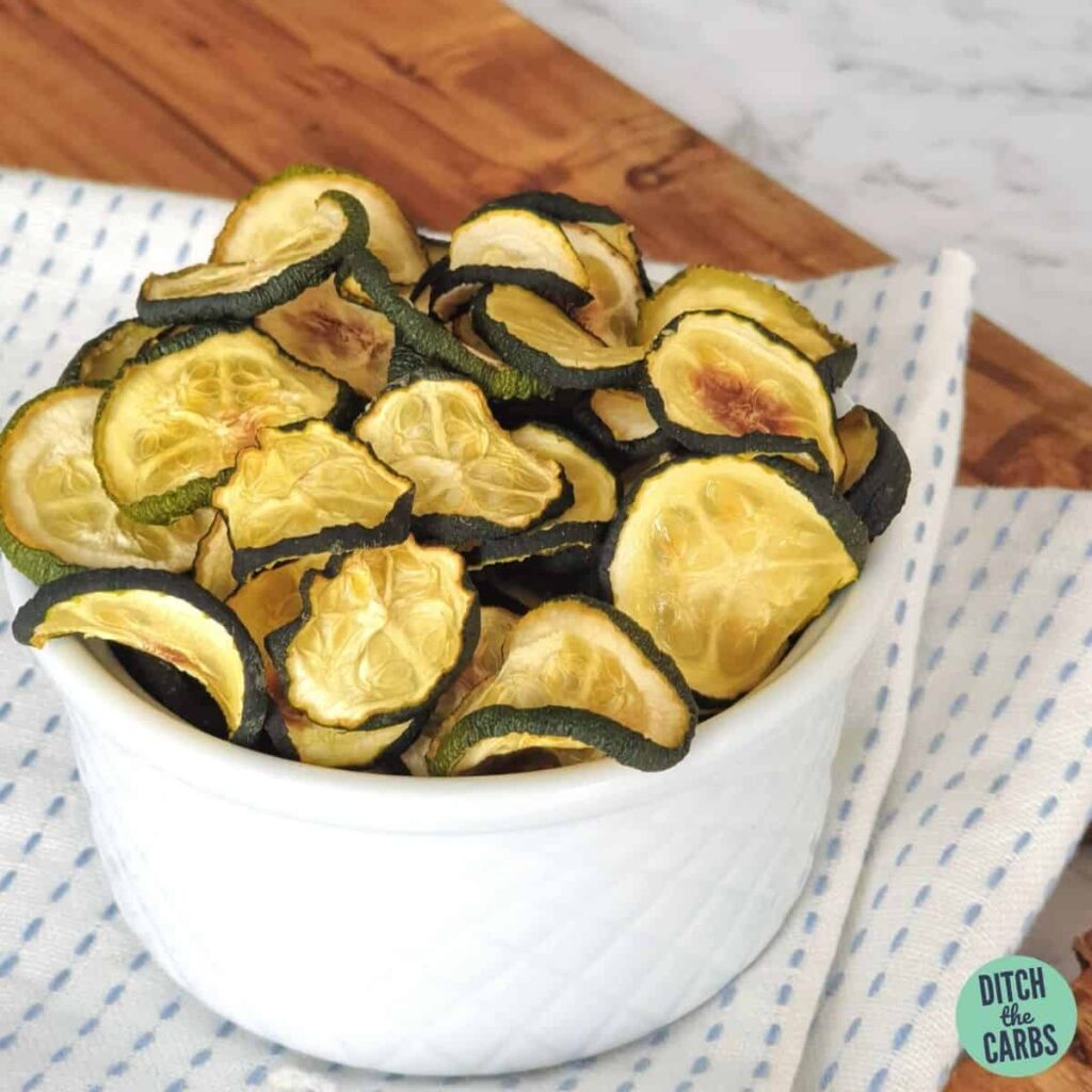 baked zucchini chips in a white bowl on a blue and white tea towel