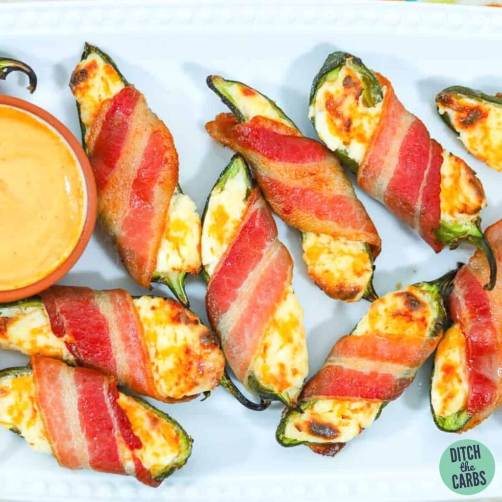 Cooked stuffed jalapeno peppers wrapped in bacon on a serving tray with a dish of spicy mayo dipping sauce.