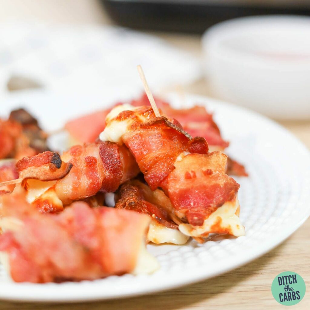 Bacon-wrapped mozzarella sticks on a white plate with a dish of marinara sauce.