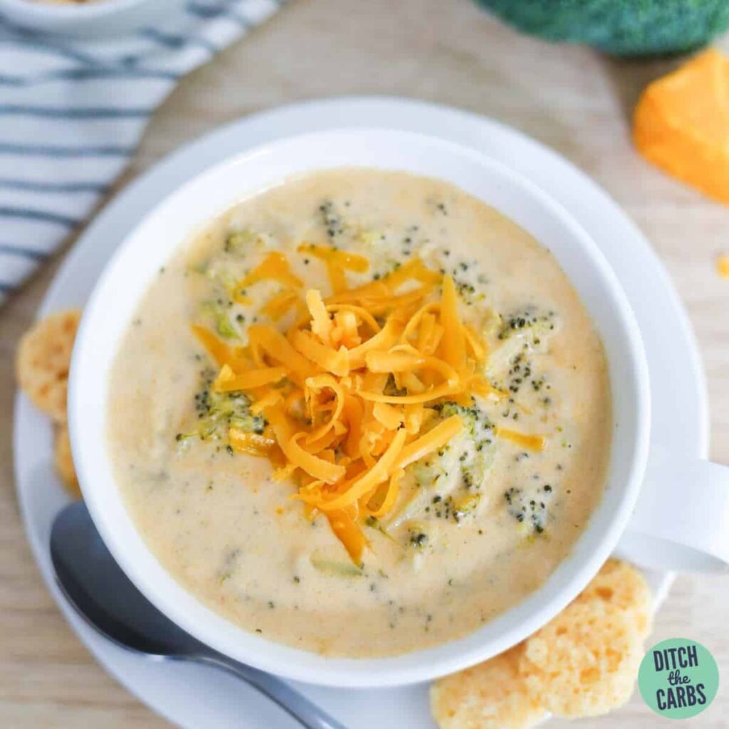 Keto broccoli cheese soup in a white soup bowl topped with shredded cheese and a side of keto crackers.