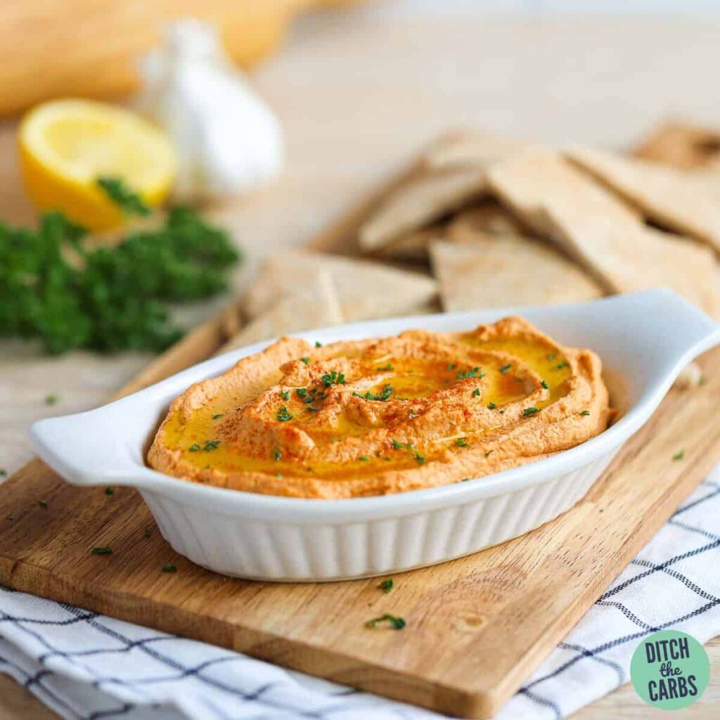 Roasted cauliflower low-carb hummus in a serving dish topped with parsley. It is resting a wooden tray with low-carb pita bread in the back for dipping.