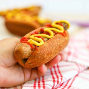A hand is holding a keto hot bun with a hot dog in it topped with yellow mustard and sugar-free ketchup.